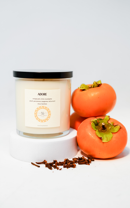 The “ADORE” candle by 55 Secret Street Candles, displayed alongside fresh persimmons and scattered cloves, emphasizing its natural and warm scent profile. The candle sits on a white pedestal with a sleek black lid, styled in a bright and clean setup.