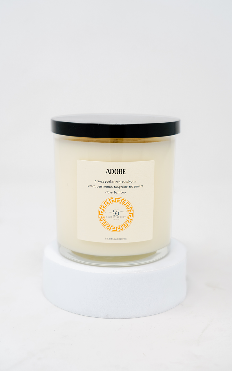 A minimalist white candle labeled “ADORE” by 55 Secret Street Candles, featuring scent notes like orange peel, citron, eucalyptus, peach, persimmon, and more. The candle is in a glass jar with a black lid, placed on a clean, white pedestal against a plain background.